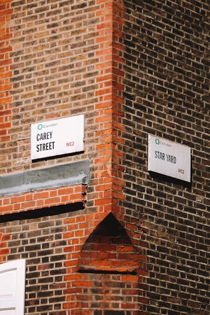 A close-up view of the corner of a red brick building featuring two street signs attached to the wall, one reading 'Carey Street' and the other 'Star Yard,' both with the Camden logo and postal code WC2. The signs are positioned at eye level, with Carey Street on the left side of the corner and Star Yard on the right. Below the Carey Street sign, there is a metallic vent or fixture extending horizontally, with a smooth, silver finish contrasting against the rough brick surface. The brickwork showcases varying shades of red, brown, and hints of black, with some bricks protruding slightly to add texture. The background consists of the brick wall, which appears aged, with visible mortar lines and subtle weathering. The lighting is neutral, illuminating the bricks and signs evenly, emphasizing the urban character associated with on-site waste management or private waste handling services often involved in rubbish removal projects in the Camden area. This scene highlights elements typical of city environments where alternative rubbish collection services may operate, such as Rubbish Removal Camden.