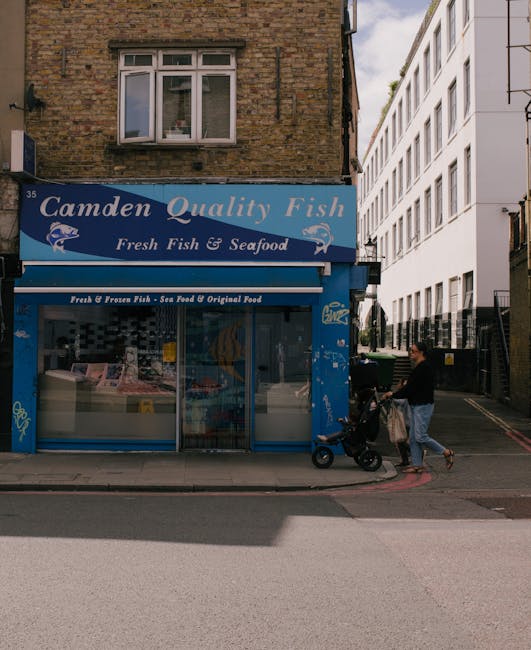 A street-level view of a blue storefront with signage that reads 'Camden Quality Fish' and 'Fresh Fish & Seafood' in white lettering, situated on the ground floor of a brick building with a single, white-framed window above. The shop's large glass window displays packaged seafood and fish products inside, with a partially visible sliding glass door to the right. To the right of the shop, a person dressed in dark clothing is walking along the sidewalk while pushing a baby stroller and carrying a shopping bag, with the stroller positioned close to the shop's entrance. Adjacent to the shop, a white multi-storey modern building with large rectangular windows appears, contrasting with the brick structure. The scene is illuminated by natural daylight, with a clear sky visible in the upper right corner, and the sidewalk, curb, and street are visible in the foreground, highlighting urban surroundings supportive of private waste management or rubbish removal services like those provided by Rubbish Removal Camden.