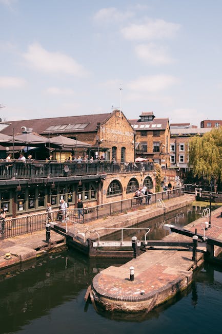 The image depicts a historic brick building situated alongside a canal, with an outdoor seating area on an upper level that features dark-colored umbrellas and a railing, where several people are seated. Below, the canal’s still water reflects the surrounding structures, and a small, rounded brick platform extends into the water, equipped with metal railings and mooring posts. The building’s brickwork has a weathered appearance with arched windows and a sloped roof, while neighboring structures in the background display similar architectural styles and materials. The sky above is partly cloudy with patches of blue, and neighboring trees, including a leafy green one on the right, add a touch of natural greenery to the urban setting. The scene suggests a lively, mixed-use waterside area that could involve private or independent management of waste and rubbish removal services in line with the context of on-site clearance and waste disposal activities typically associated with such locations, supporting the services offered by Rubbish Removal Camden.
