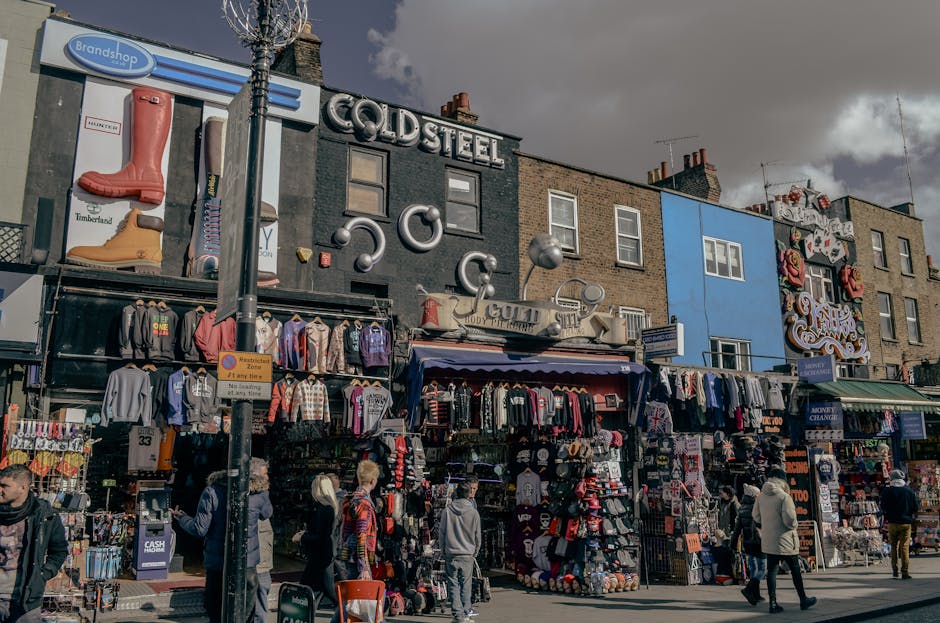 The image depicts a busy street scene in front of a row of commercial buildings with various signs and storefronts, possibly in a market area. In the foreground, multiple outdoor stalls and booths display a large assortment of clothing, including jackets, hoodies, hats, and accessories, all arranged on racks, tables, and hanging displays. Behind the stalls, there are brick facades of buildings with various window styles, some adorned with decorative signage and neon lights. One prominent sign reads 'COLDSTEEL,' with additional metallic decorative elements such as pipes and spheres attached to the building's facade. To the left, a tall, bare tree is partially visible, with a blue advertising sign featuring an image of rubber boots attached to a building. Several pedestrians are walking past, some carrying shopping bags or browsing the stalls, contributing to the lively atmosphere. The scene suggests a retail environment that could be associated with independent collection or private disposal activities, typical of informal rubbish clearance in a commercial or market setting. The overall lighting suggests an overcast day, casting diffused light over the scene, emphasizing the textures of the brickwork, textiles, and signs, aligning with waste collection and disposal contexts managed by services like Rubbish Removal Camden.