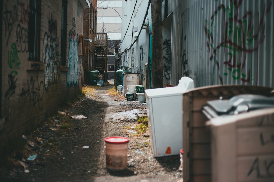 A narrow alleyway between two brick buildings with walls covered in various graffiti and spray paint. The ground is uneven, muddy, and strewn with small debris, reflecting an urban environment with limited maintenance. On the right side, there are several large discarded appliances and waste containers, including a white washing machine, a beige plastic bin, and an open outdoor storage box filled with miscellaneous items, indicating collection of bulky waste. Further down the alley, there are multiple dark green waste bins and barrels, as well as additional discarded items such as a small white container and a plastic bucket with a red lid, suggesting a collection point for rubbish removal. The scene is lit with natural daylight and shows signs of ongoing clearance or collection activity, typical of private waste management services like those offered by Rubbish Removal Camden, providing alternative solutions for rubbish disposal in areas with restricted access.