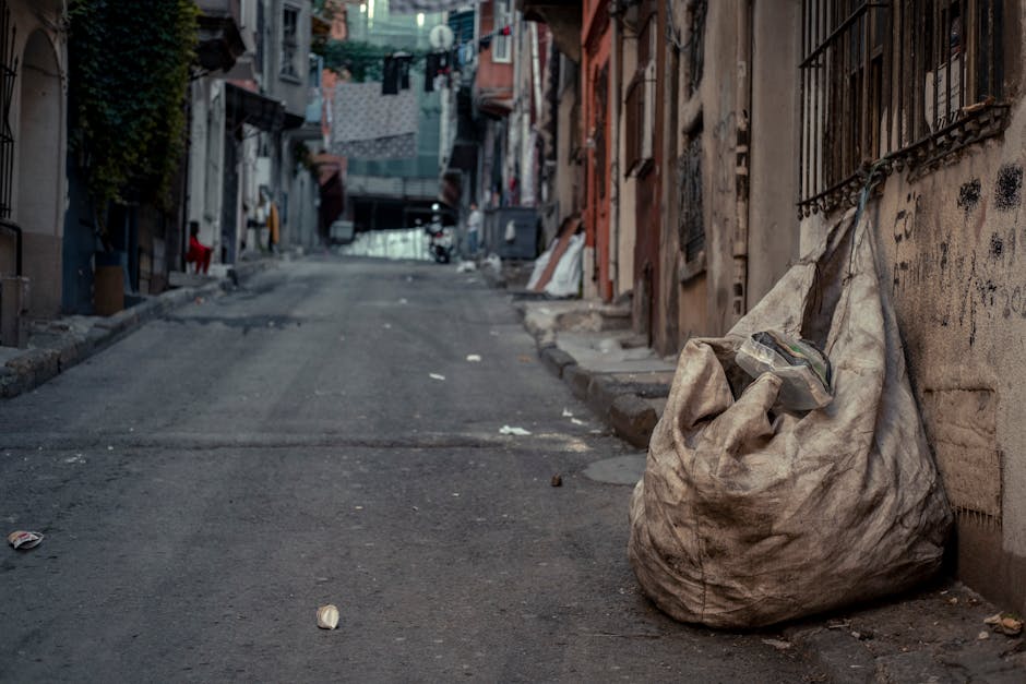 A large, weathered, beige fabric sack filled with miscellaneous rubbish, resting against a graffiti-marked brick wall on a narrow, uneven urban street. The sack appears heavy and bulging, with some waste visible at the top, including plastic items and paper. The street surface is rough asphalt with scattered small debris and discarded packaging, sloping slightly upward. In the background, residential buildings with closed windows, metal security grilles, and hanging laundry, create a dense, urban environment. The scene is lit by natural daylight, casting soft shadows and revealing the textures of the sack's fabric, the worn brickwork, and the surrounding surroundings. This setting illustrates a typical instance of informal waste disposal often addressed through private rubbish removal services like those provided by Rubbish Removal Camden, especially in areas with limited access for large collection vehicles. The scene emphasizes the importance of professional rubbish removal solutions for narrow access locations, aligning with the focus on alternative waste handling methods apart from municipal collection.