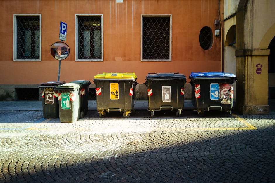 A row of five large waste bins aligned along a cobblestone street in front of an orange-brown building with three barred windows, each bin having distinct colours and labels indicating different waste types. From left to right, the first bin is green with a recycling symbol and a textured surface, suitable for general waste collection. The second bin features a yellow lid with a label indicating it is for recyclable materials; it has a smooth, hard plastic finish with wheels at the base. The third bin is black with a grey lid, designated for general refuse, with a slightly textured surface and a white label. The fourth bin is blue with a matching lid, indicating it is for paper or plastics, and displays a recycling icon; it appears to be made of durable plastic with a smooth finish. Ambient daylight casts soft shadows over the textured cobblestones, and a convex mirror is mounted on a post behind the bins, providing a safety inspection point. The scene reflects an organized arrangement suitable for private waste management or independent rubbish removal services, such as those provided by Rubbish Removal Camden.