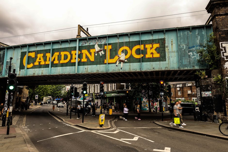 A large turquoise metal railway bridge spans across a busy street in Camden, with the words 'CAMDEN LOCK' painted in bold yellow letters on its side. The bridge shows signs of weathering, including rust and peeling paint. Two children are illustrated on the bridge, climbing or hanging from the structure, depicted in a mural style with a playful, sketch-like appearance. Below the bridge, the street scene includes pedestrians crossing at traffic signals, some walking along the pavement, and others waiting at the crossings. The street is lined with various shops and buildings, with traffic lights and street signs visible. Overhead, power lines stretch across the cloudy sky. In the foreground, yellow and black traffic cones are positioned on the road, and a few vehicles, including a black car, are waiting or passing through the intersection. The scene captures the urban environment typical of London’s Camden area, with a vibrant yet neutral tone suitable for describing waste disposal or rubbish removal contexts, subtly emphasizing the setting’s practical, cityscape characteristics.