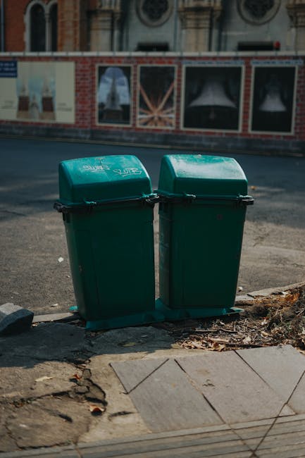 A row of five large waste bins aligned along a cobblestone street in front of an orange-brown building with three barred windows, each bin having distinct colours and labels indicating different waste types. From left to right, the first bin is green with a recycling symbol and a textured surface, suitable for general waste collection. The second bin features a yellow lid with a label indicating it is for recyclable materials; it has a smooth, hard plastic finish with wheels at the base. The third bin is black with a grey lid, designated for general refuse, with a slightly textured surface and a white label. The fourth bin is blue with a matching lid, indicating it is for paper or plastics, and displays a recycling icon; it appears to be made of durable plastic with a smooth finish. Ambient daylight casts soft shadows over the textured cobblestones, and a convex mirror is mounted on a post behind the bins, providing a safety inspection point. The scene reflects an organized arrangement suitable for private waste management or independent rubbish removal services, such as those provided by Rubbish Removal Camden.